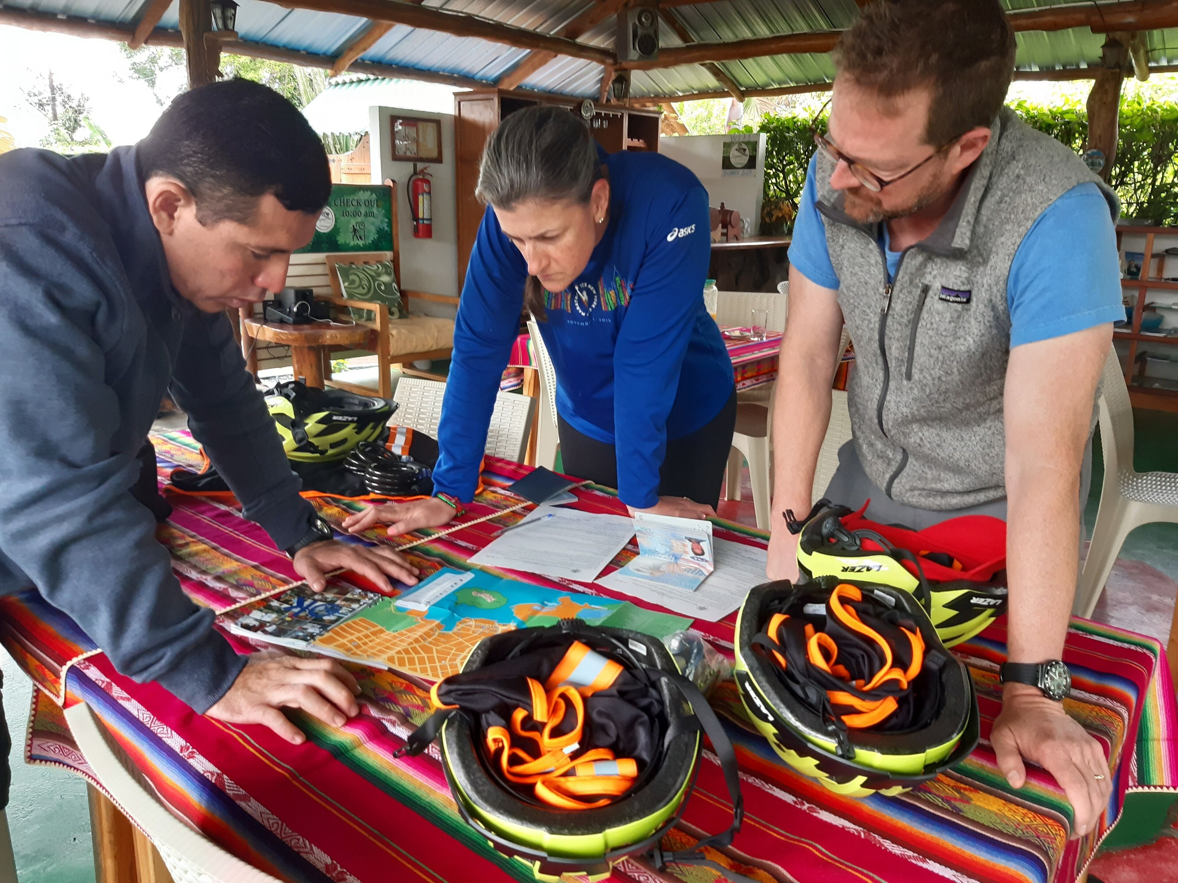 Tres personas sentadas a una mesa con mantel colorido, conversando sobre objetos que hay encima, incluyendo un mapa y una bolsa amarilla.