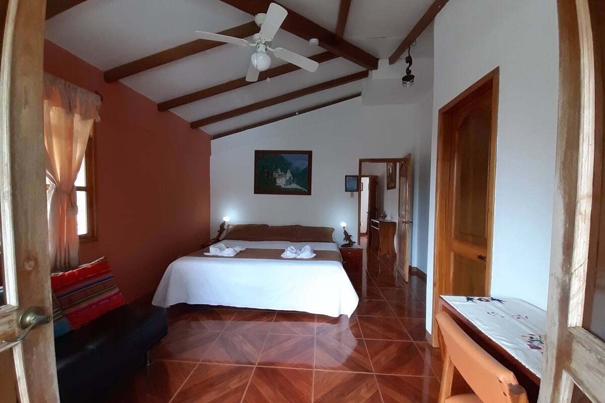 A hotel room with a bed, a desk, a chair, a ceiling fan, and a painting on the wall.