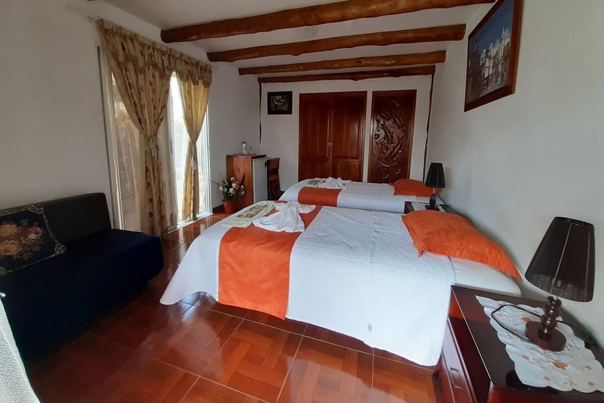 A room with a wooden ceiling, a bed with orange and white bedding, a desk with a lamp, and a chair.