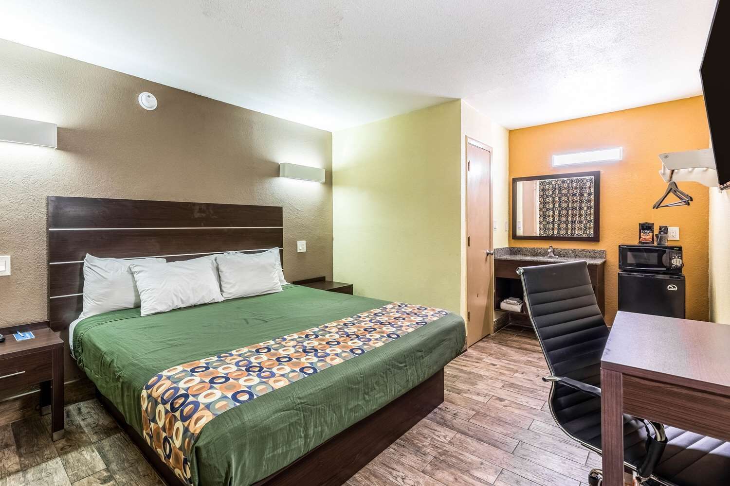 A hotel room with a bed, a desk, a chair, a microwave, and a small kitchenette.