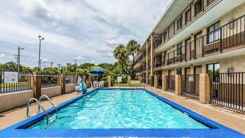 Une piscine bordée de bleu, un espace détente avec des chaises et un parasol, et un immeuble à plusieurs étages doté de balcons.