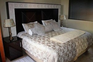 Cama bem arrumada com colcha estampada e almofadas, com duas luminárias laterais sobre mesas de cabeceira escuras.