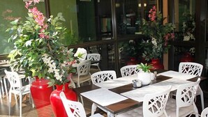 Un restaurant avec des chaises et des tables blanches, de grands vases rouges ornés de fleurs et des plantes en pot.