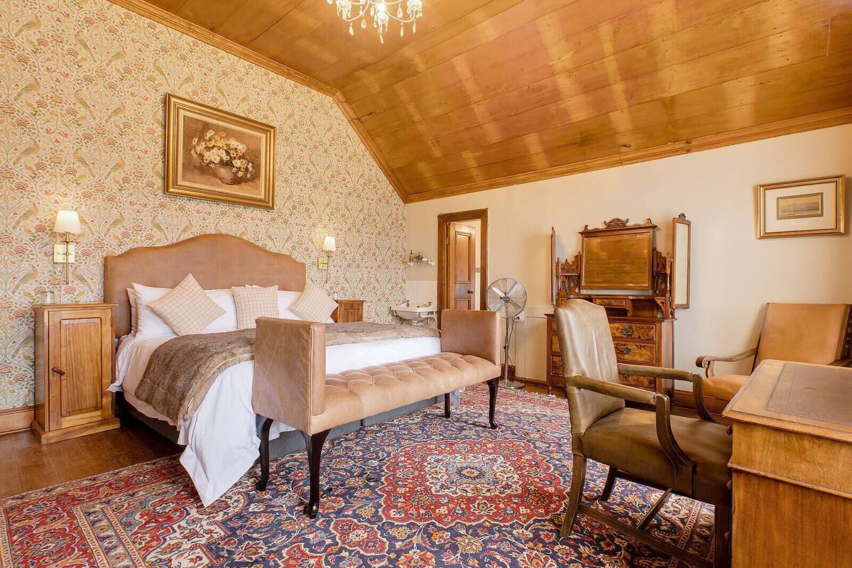 Une chambre à coucher avec un grand lit, une tête de lit en bois, un banc, un bureau, une chaise, un ventilateur et un miroir.