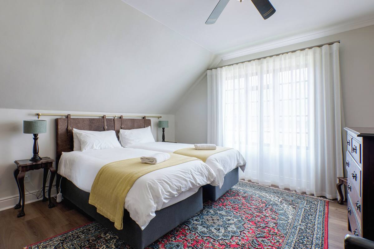 Un lit double avec une tête de lit en bois, une table de chevet avec une lampe, une grande fenêtre avec des rideaux légers et un tapis à motifs.