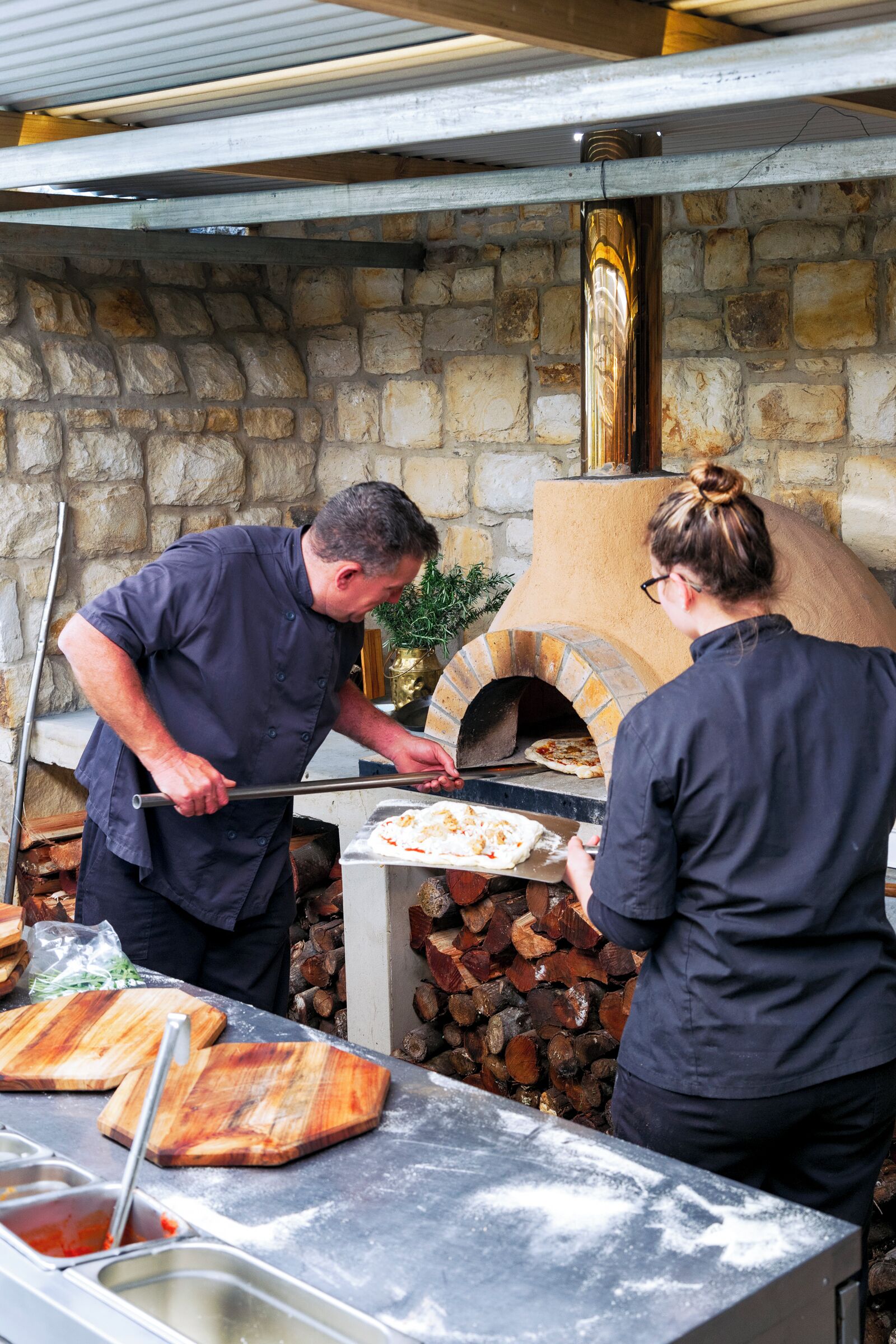 A stone wall with a brick oven and a metal counter with pizza dough and toppings.