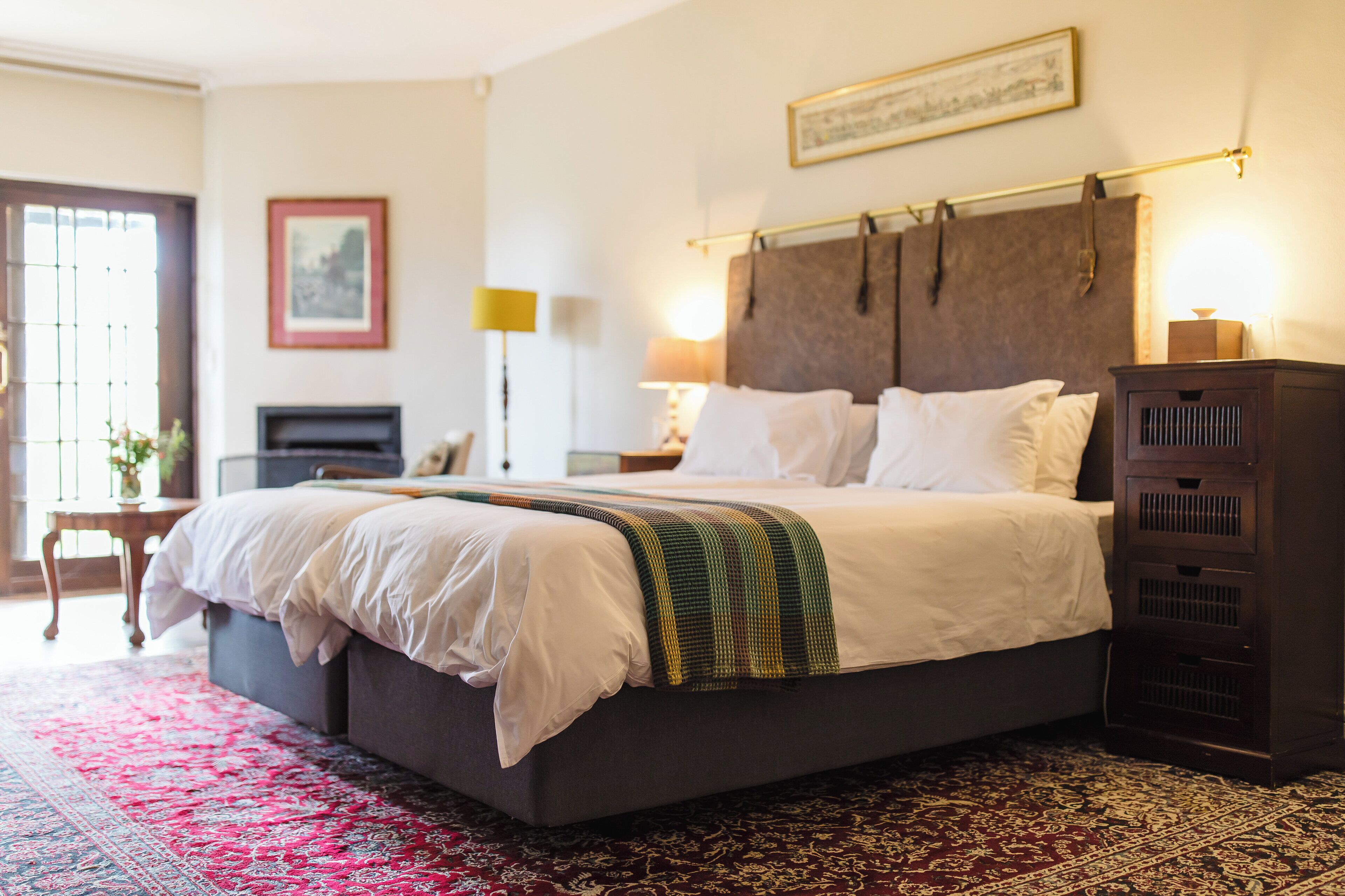 Een net opgemaakt bed met witte lakens en een decoratieve plaid, een donker houten hoofdbord en een rood, patroonrijk vloerkleed.