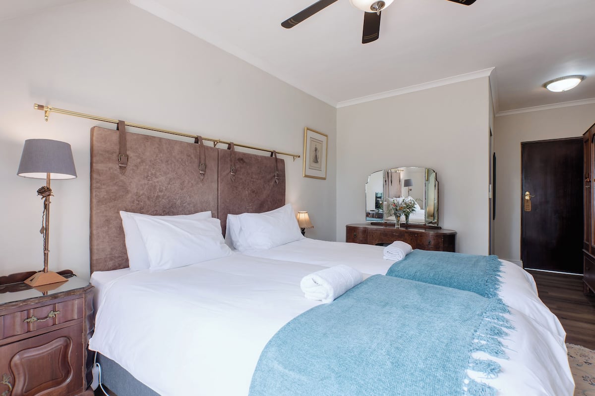 Une chambre d’hôtel avec un grand lit, une table de chevet avec une lampe, une commode, un miroir et un ventilateur de plafond.