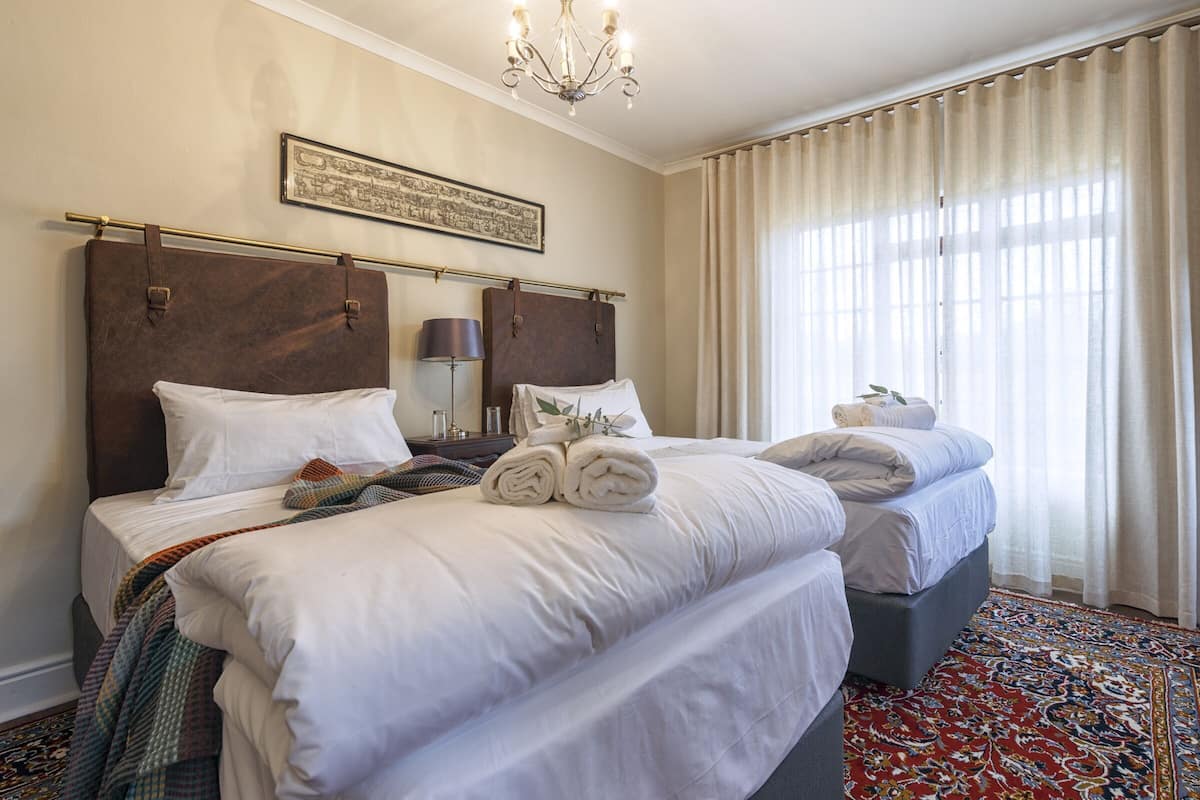 Une chambre d’hôtel avec deux lits, un lustre, un tableau encadré au mur et une grande fenêtre avec des rideaux.