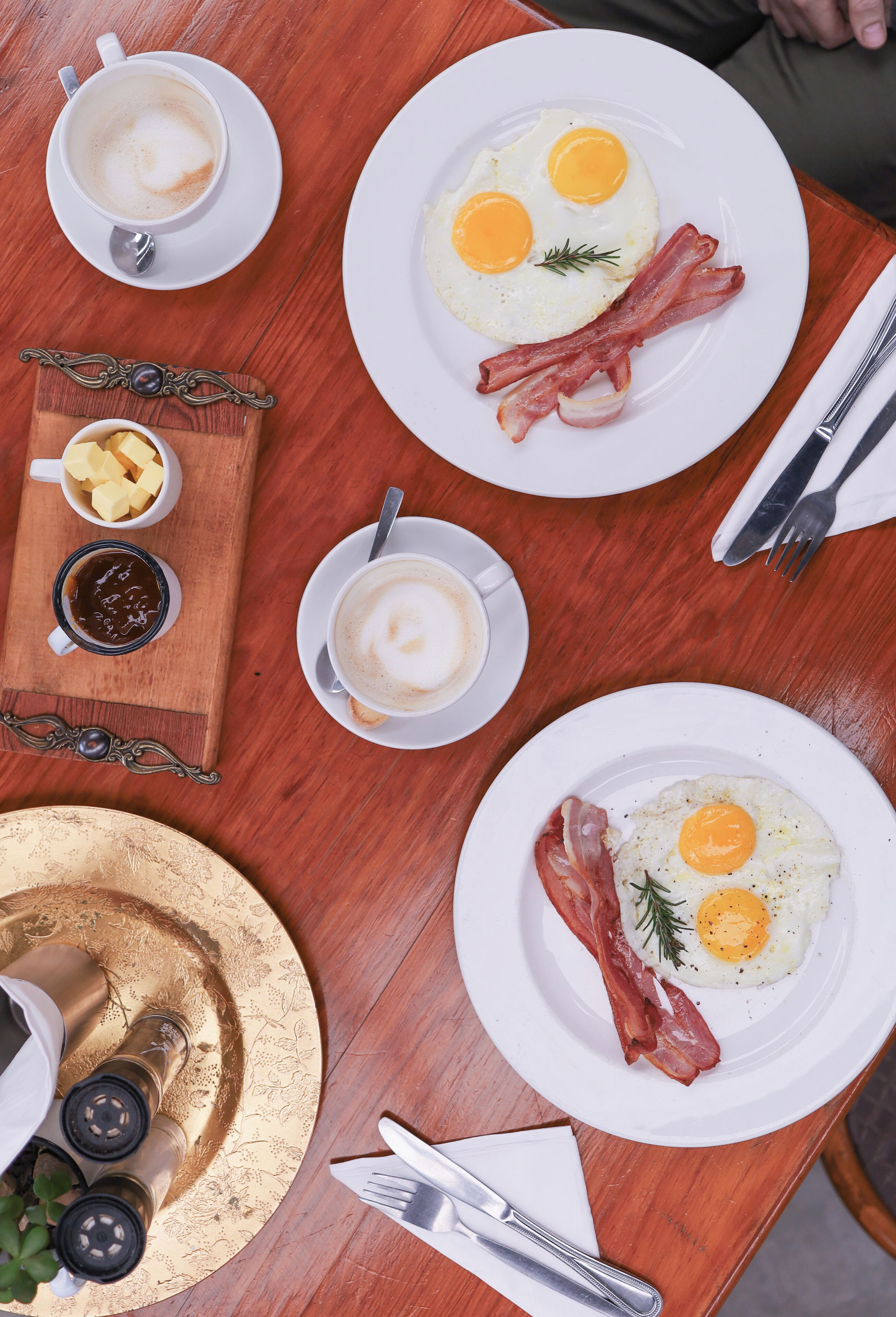A breakfast setting with plates of eggs and bacon, coffee cups, and a wooden board with butter and jam.