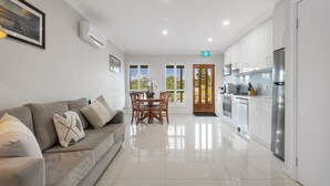 A modern kitchen with white cabinets and stainless steel appliances, a dining area with a wooden table and chairs, and a living area with a sofa and cushions.