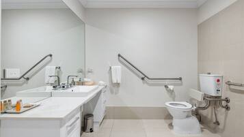 Une salle de bain moderne avec un lavabo, un miroir et des toilettes blancs. Elle est équipée de barres d'appui et d'une poubelle.