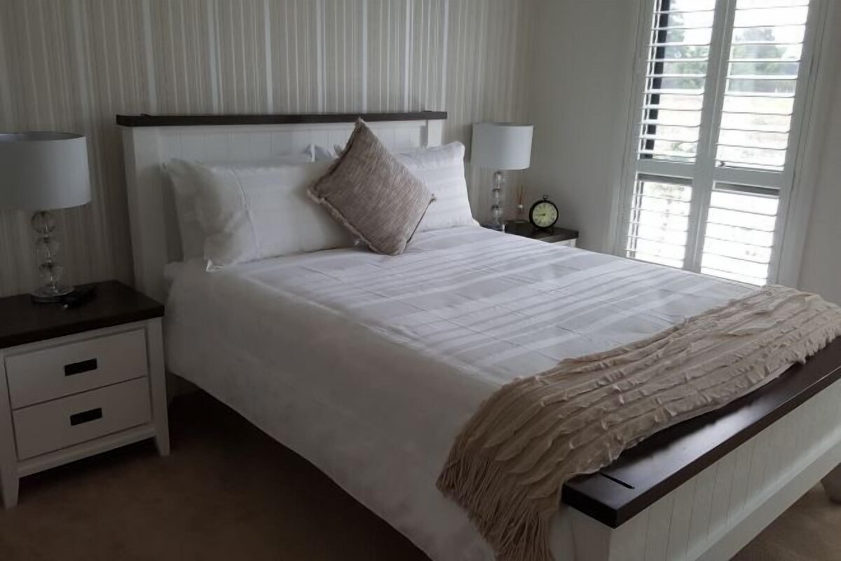 Un lit bien fait, recouvert d’une couette blanche et d’un plaid beige, avec deux tables de chevet de chaque côté, sur lesquelles sont posées des lampes et une horloge. La pièce est décorée d’un papier peint à rayures verticales et possède une fenêtre avec des stores.