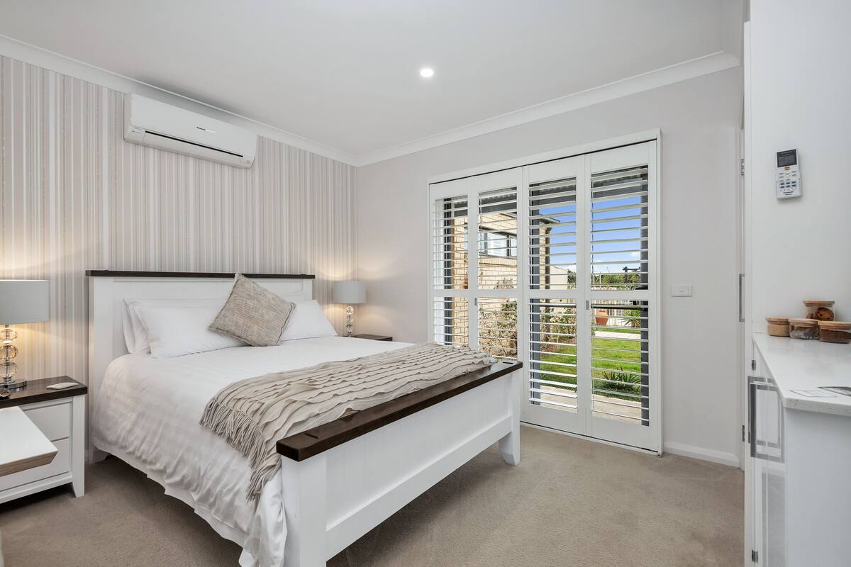 A bedroom with a bed, bedside tables, a nightstand, a window with blinds, and an air conditioning unit.