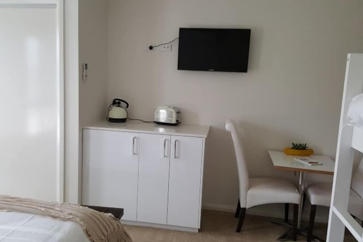 A bedroom with a bed, a white wardrobe, a desk with a chair, a small table with a plant, and a TV mounted on the wall.