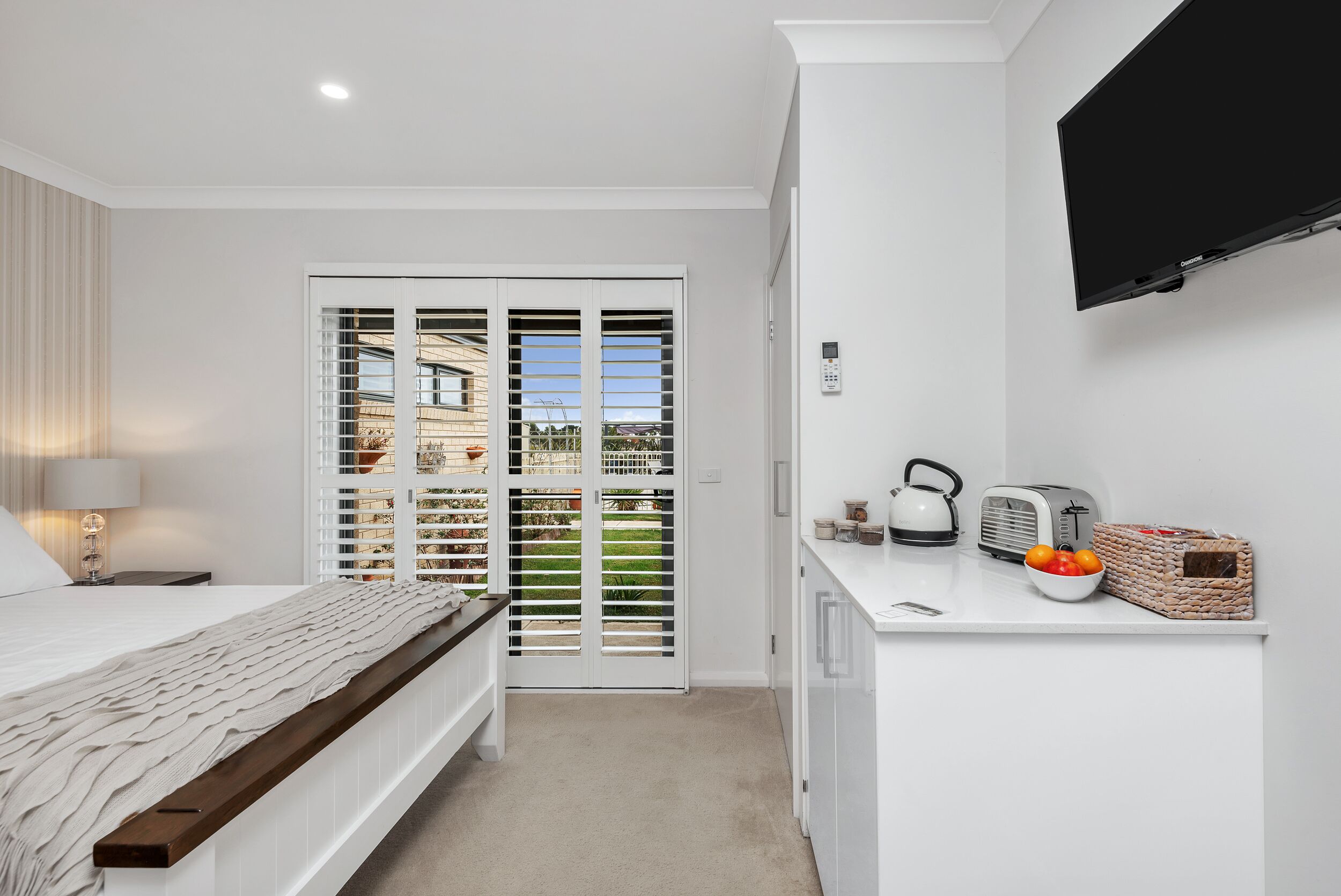A bedroom with a bed, a nightstand, a lamp, a TV, a kitchen area with a toaster, kettle, and fruit bowl.