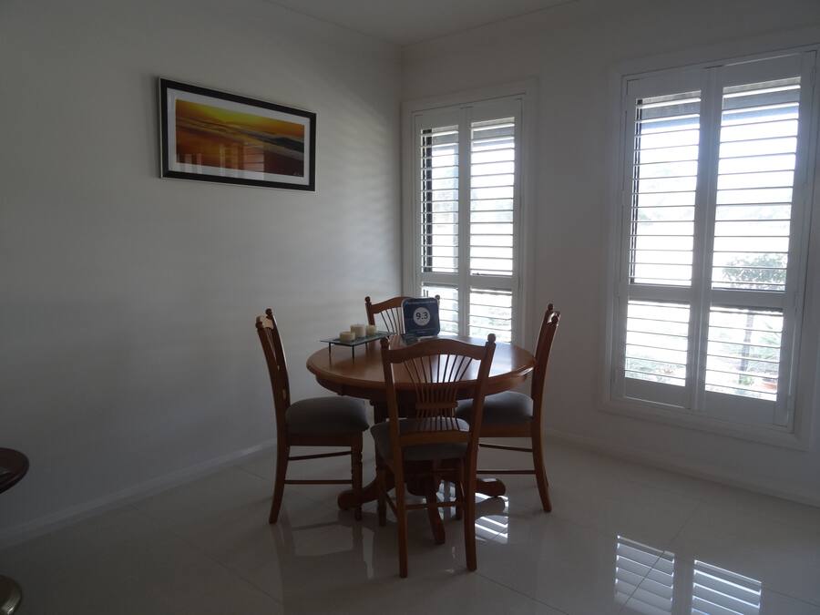 A dining area with a round table and four chairs, a framed picture on the wall, and large windows with blinds.