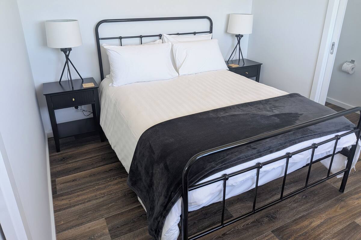 A neatly made bed with a black metal frame, white bedding, and a black throw blanket. There are two bedside tables with lamps, a small drawer, and a wooden nightstand with a lamp. The room has wooden flooring and light gray walls.