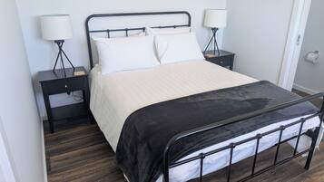 A neatly made bed with a black metal frame, white bedding, and a black throw blanket. There are two bedside tables with lamps, a small drawer, and a wooden nightstand with a lamp. The room has wooden flooring and light gray walls.