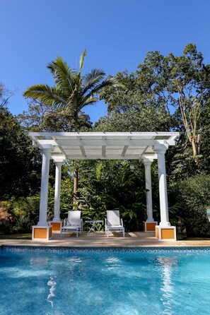 A pool area with a pergola, white chairs, and lush greenery in the background. - Locanda Della Mimosa (Petrópolis)