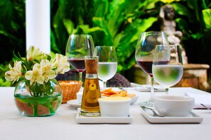 Una mesa dispuesta con copas de vino, un bol de queso y una botella de aceite de oliva, rodeada de follaje y una estatua.