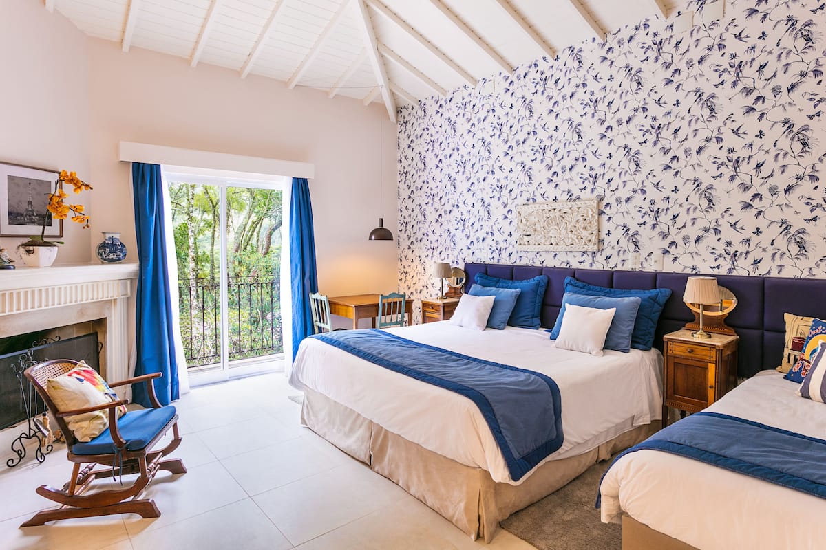 A bedroom with two beds, a fireplace, a rocking chair, and a balcony with a view of greenery.