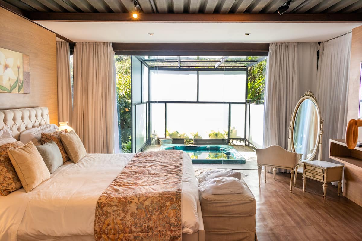 A spacious bedroom with a large bed, a sitting area, a vanity with a mirror, and a view of a swimming pool.