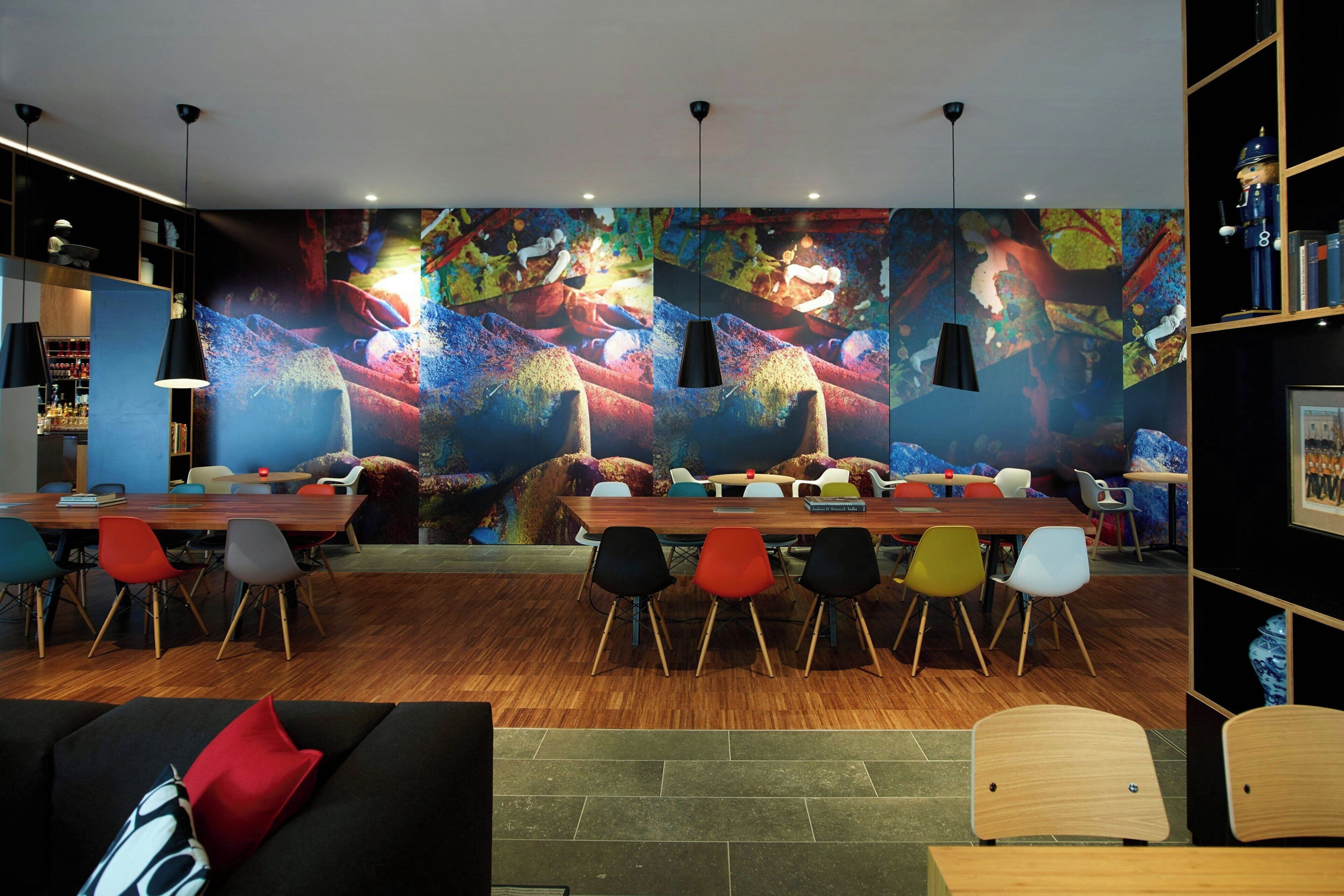 A modern dining area with a long wooden table, colorful chairs, and a vibrant abstract mural on the wall.