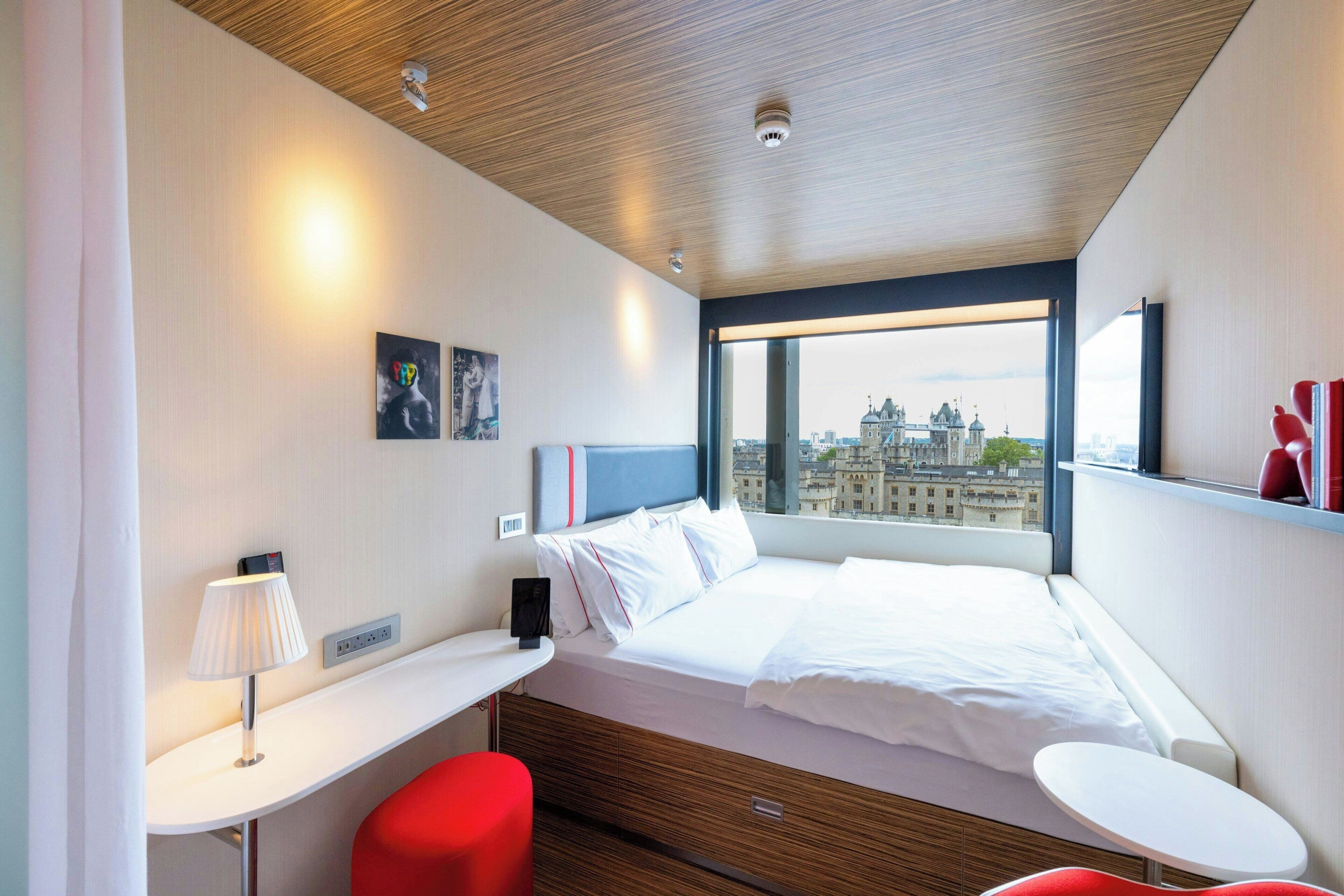 Ett modernt hotellrum med en säng, ett skrivbord, en stol och ett fönster med utsikt över staden.