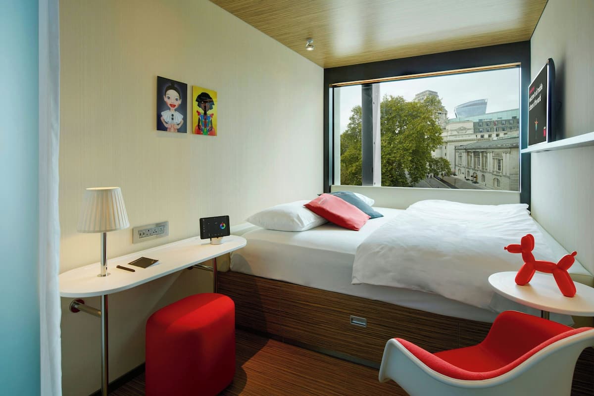 A hotel room with a bed, a desk, a chair, and a window with a view of buildings.