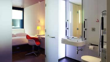Habitación de hotel con cama, una silla roja, mesita de noche con lámpara y baño con lavamanos, inodoro y ducha.