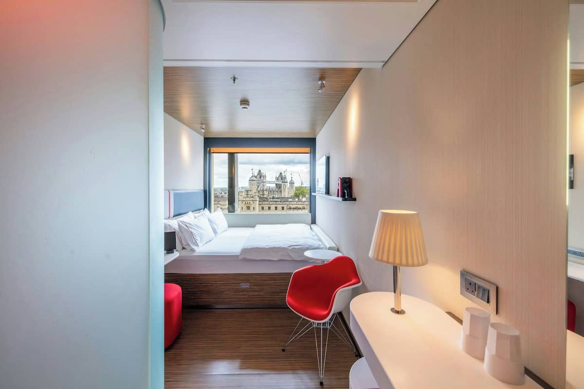 A modern hotel room with a bed, a red chair, a desk, and a lamp.