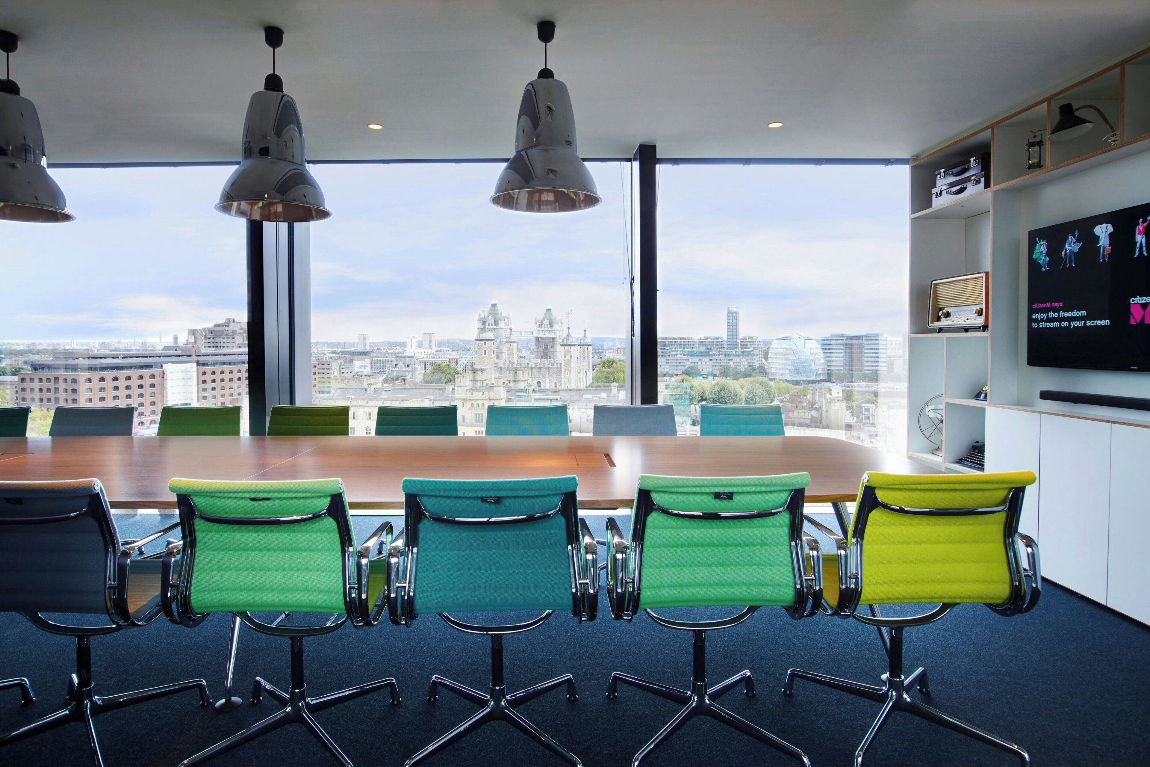 A conference room with a large table and green chairs, a city view, and a television on the wall.