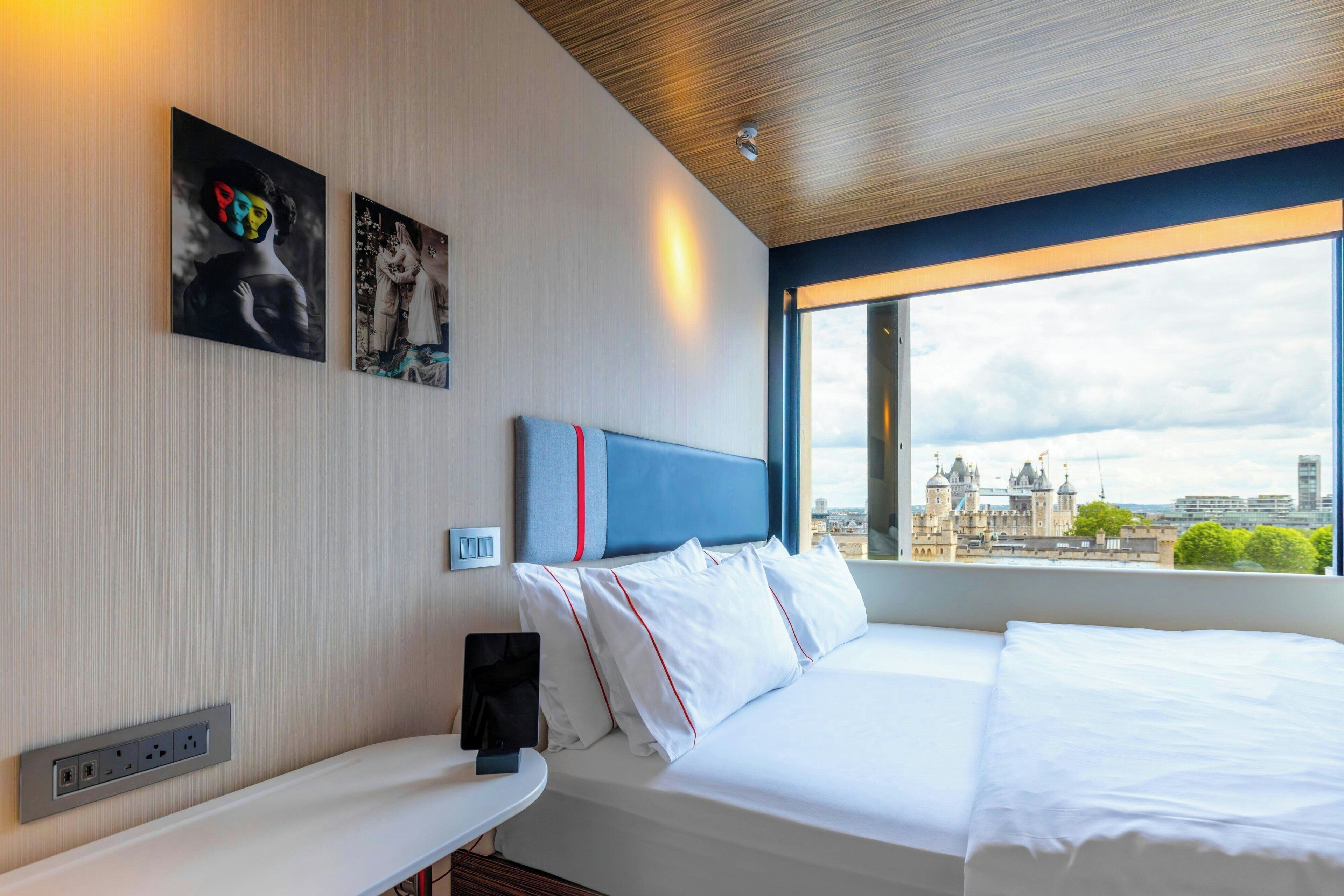 Ein Hotelzimmer mit Bett, Nachttisch, Wandsteckdose und Fenster mit Blick auf die Stadt.