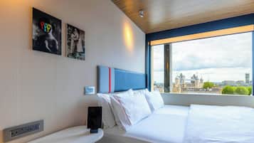 Ein Hotelzimmer mit Bett, Nachttisch, Wandsteckdose und Fenster mit Blick auf die Stadt.