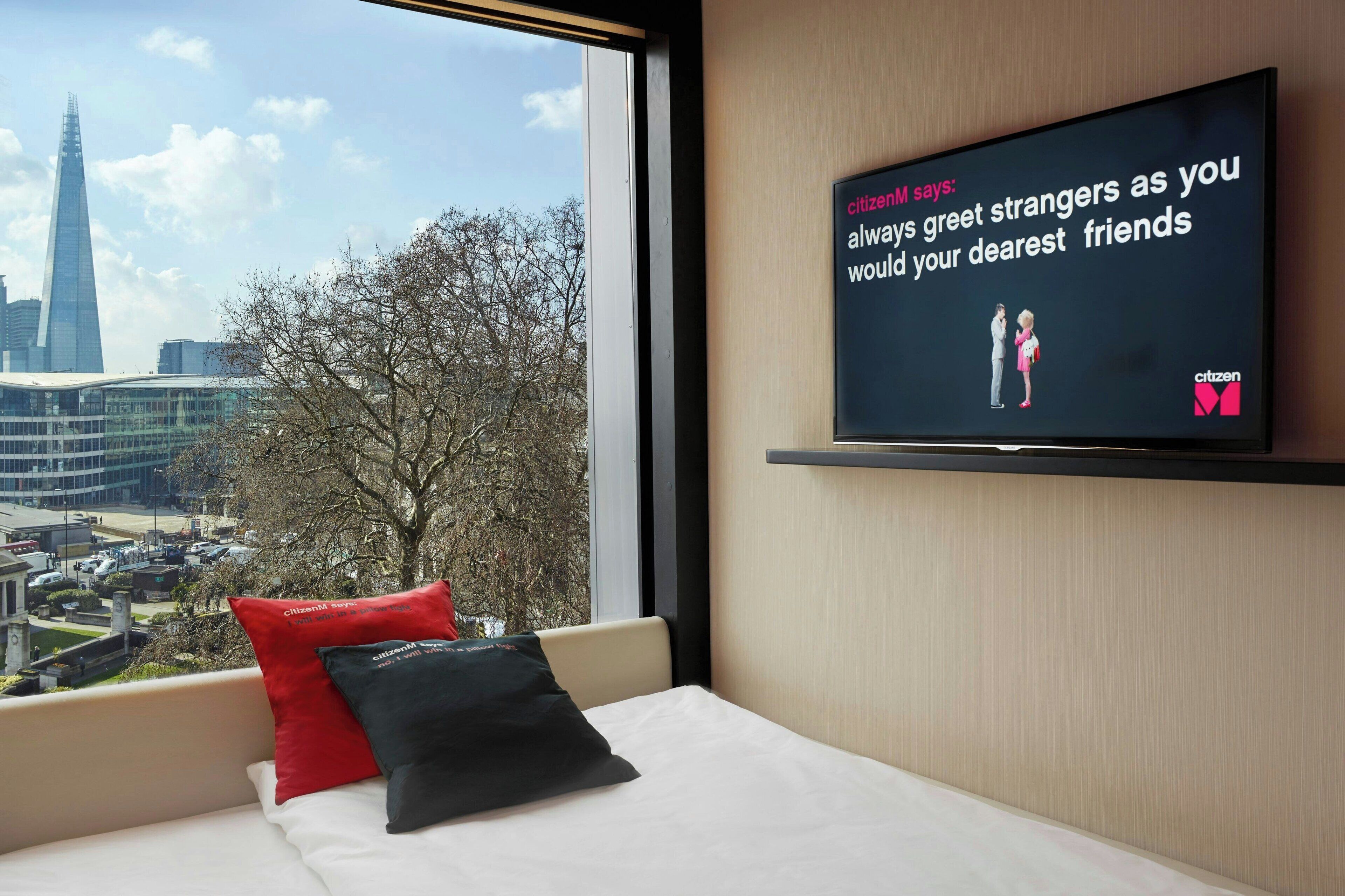 A hotel room with a large window, a bed with white bedding, two pillows, and a television displaying a message.