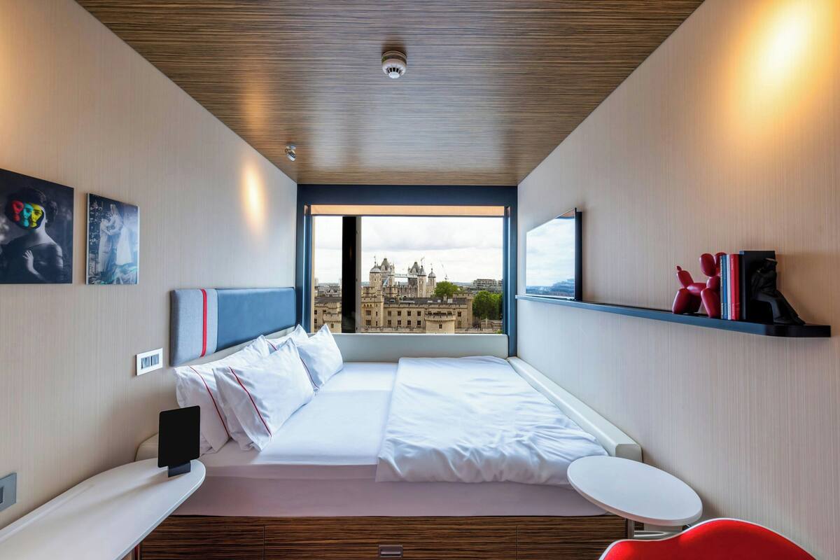 A modern hotel room with a large bed, a small table, and a view of a cityscape through a large window.