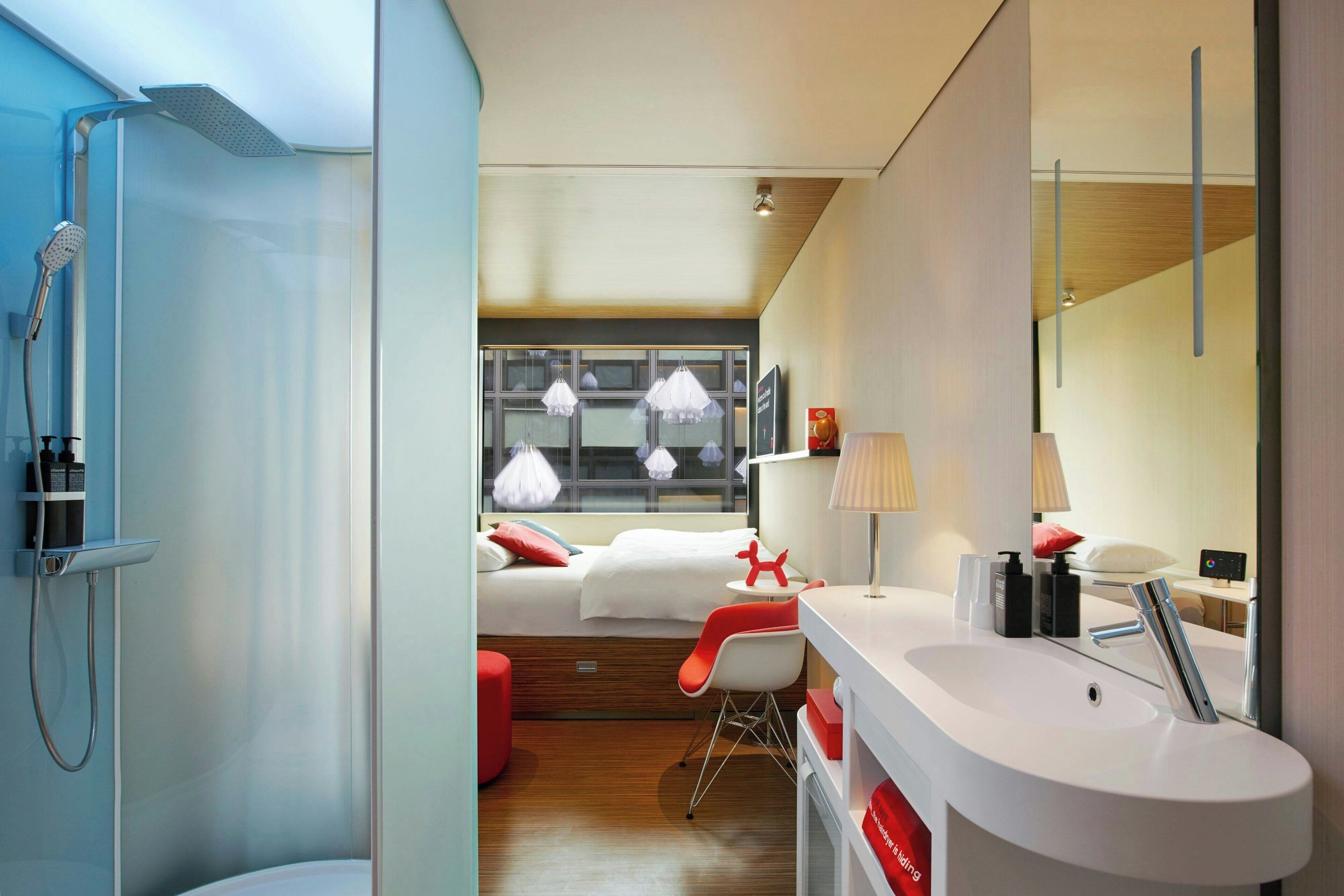 A modern hotel room with a shower, a bed with white bedding, a red chair, a white sink, and a large window with hanging lights.