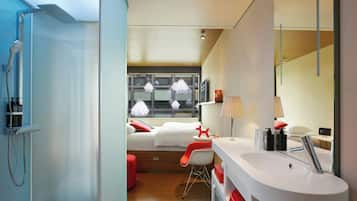 A modern hotel room with a shower, a bed with white bedding, a red chair, a white sink, and a large window with hanging lights.