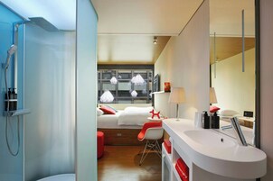 A modern hotel room with a shower, a bed with white bedding, a red chair, a white sink, and a large window with hanging lights.