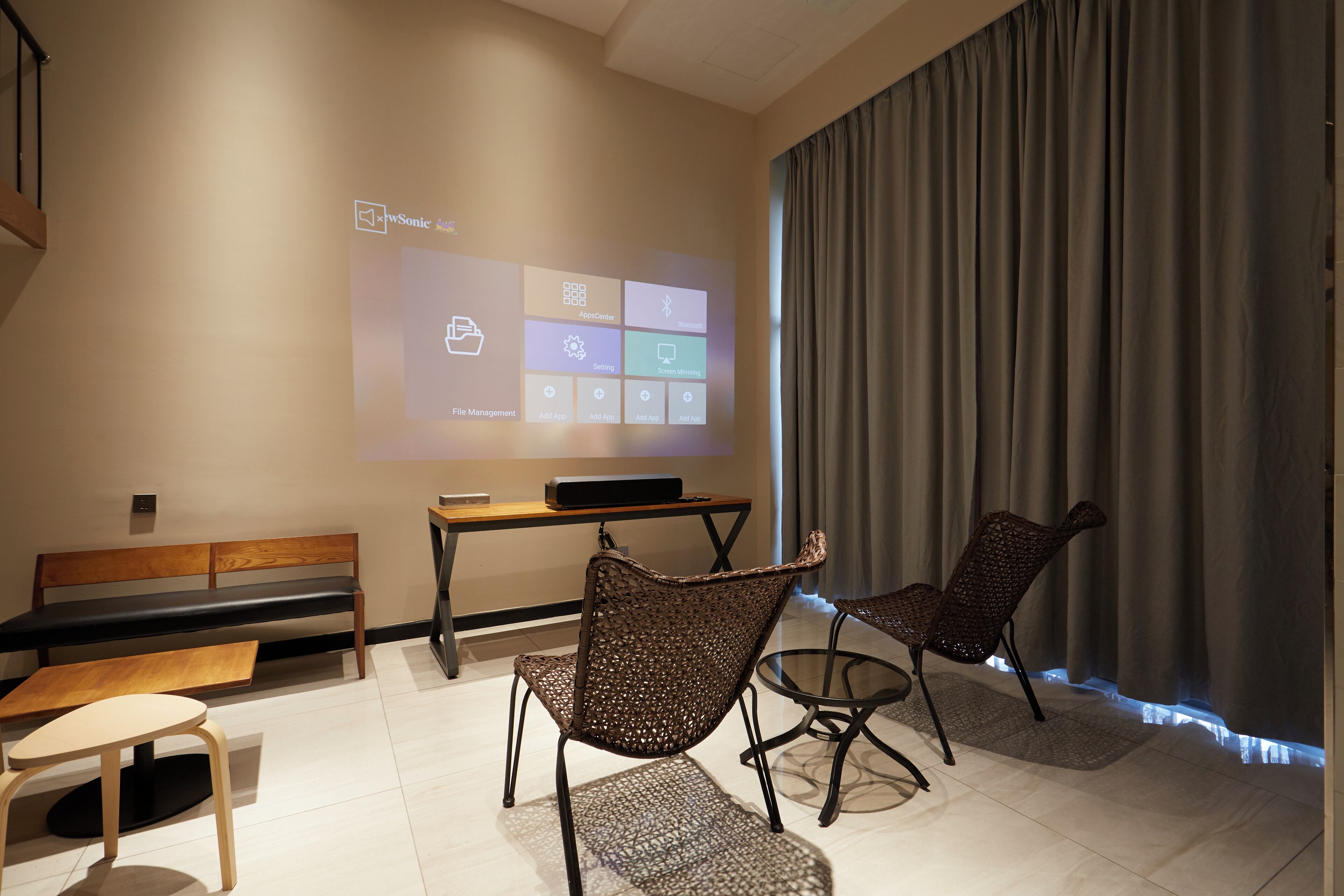 Una habitación moderna con una pantalla de proyección mostrando una interfaz de software, un banco de madera, dos sillas de mimbre y una pequeña mesa.