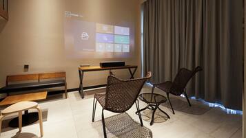 Una habitación moderna con una pantalla de proyección mostrando una interfaz de software, un banco de madera, dos sillas de mimbre y una pequeña mesa.