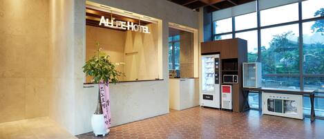 A hotel reception area with a reception desk, a potted plant, a vending machine, and large windows offering a view of the outdoors.