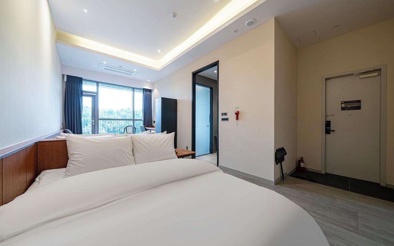 A hotel room with a large bed, a desk, a chair, a window with curtains, a door, and a fire alarm.