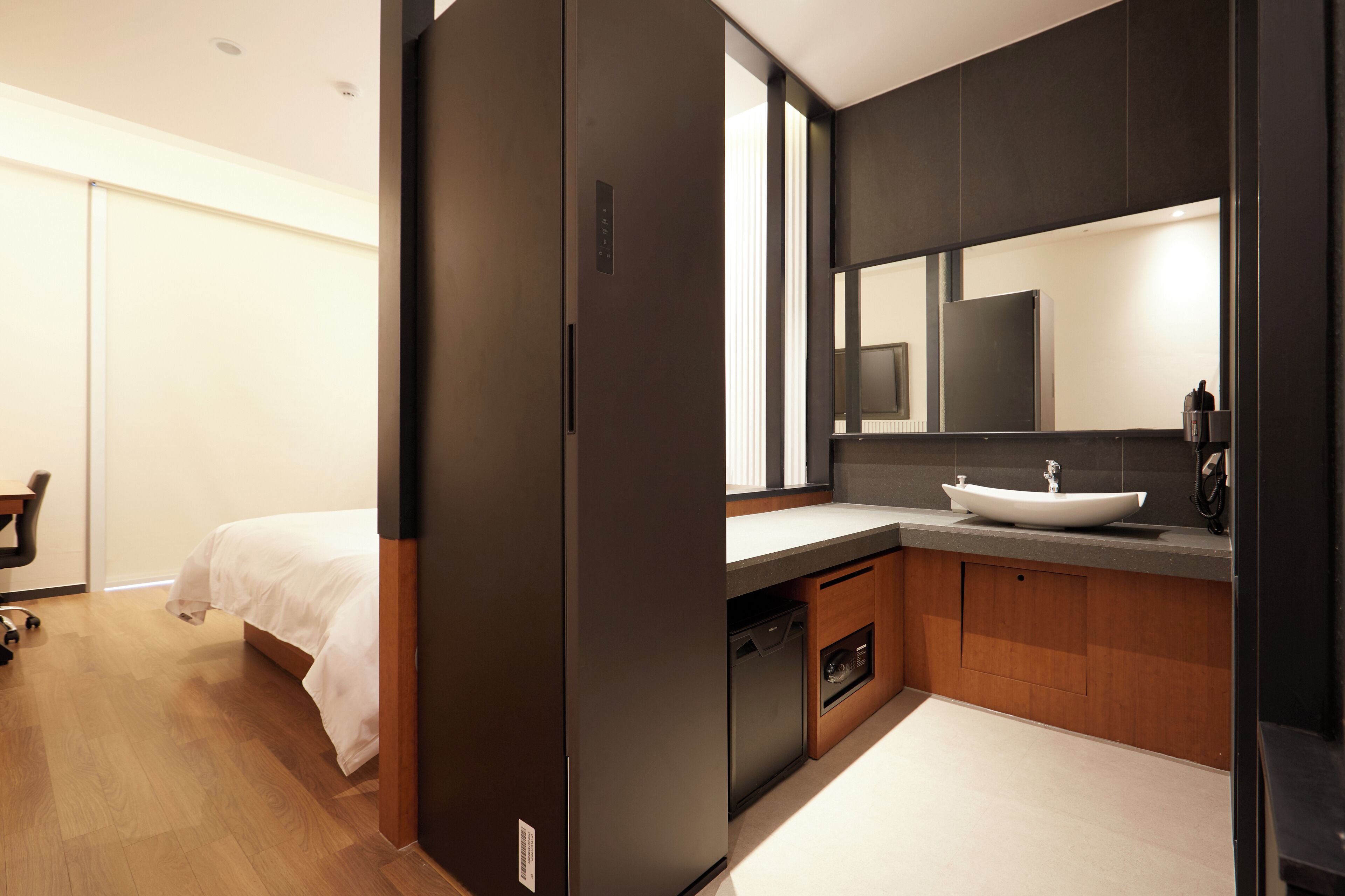Quarto de hotel moderno com piso de madeira, cama com roupa de cama branca, guarda-roupa escuro, banheiro com espelho grande, pia e vaso sanitário.