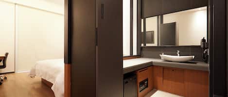 A modern hotel room with a wooden floor, a bed with white bedding, a dark-colored wardrobe, a bathroom with a large mirror, a sink, and a toilet.