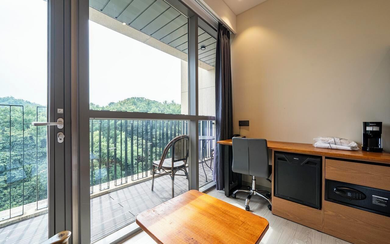 A modern room with a balcony, a wooden desk, a chair, a small table, and a coffee machine.