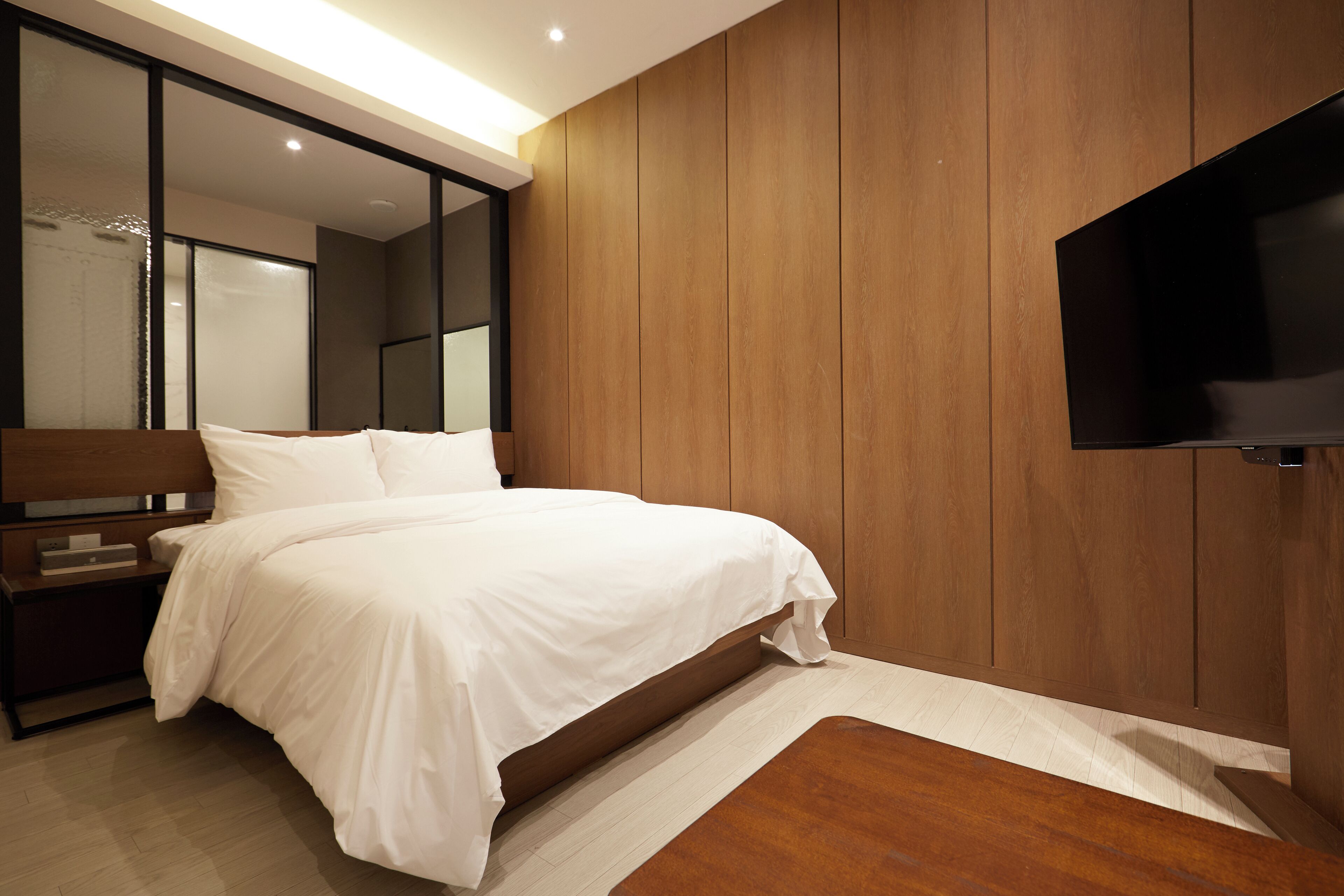 Een moderne slaapkamer met een groot bed, houten paneelwanden, een flatscreen televisie aan de muur en een nachtkastje met een lamp.