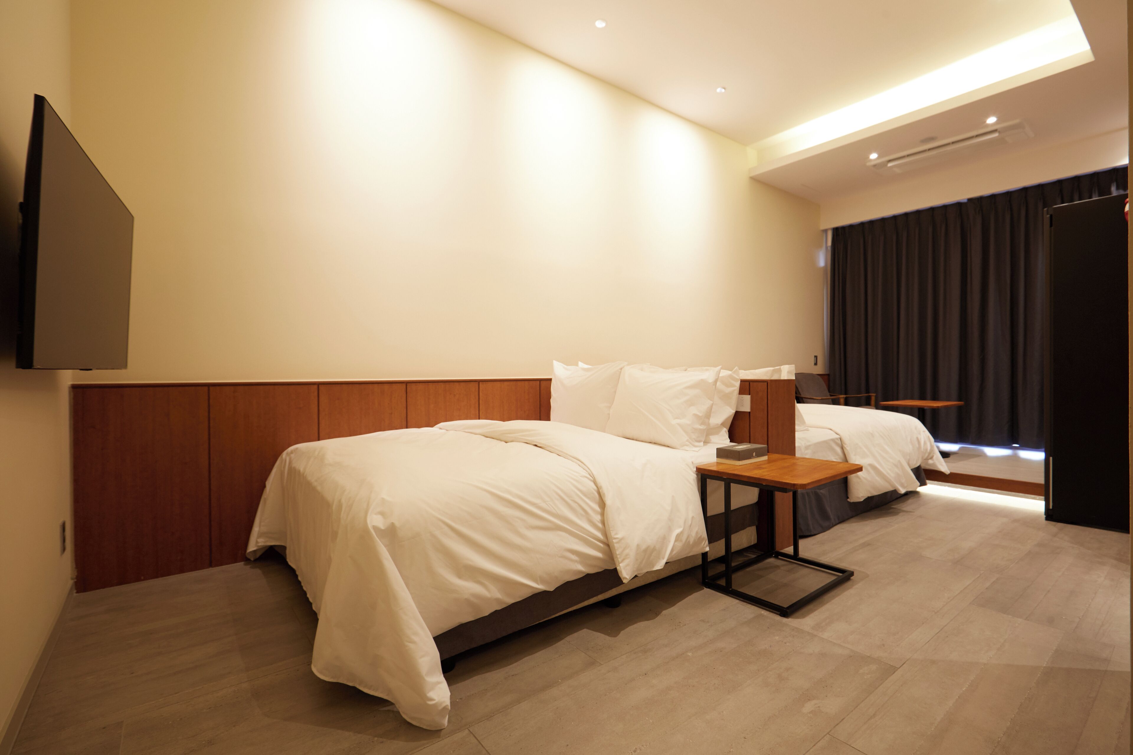 Ein Hotelzimmer mit zwei Betten, einem Flachbildfernseher, einem kleinen Tisch und einem Fenster mit Vorhängen.