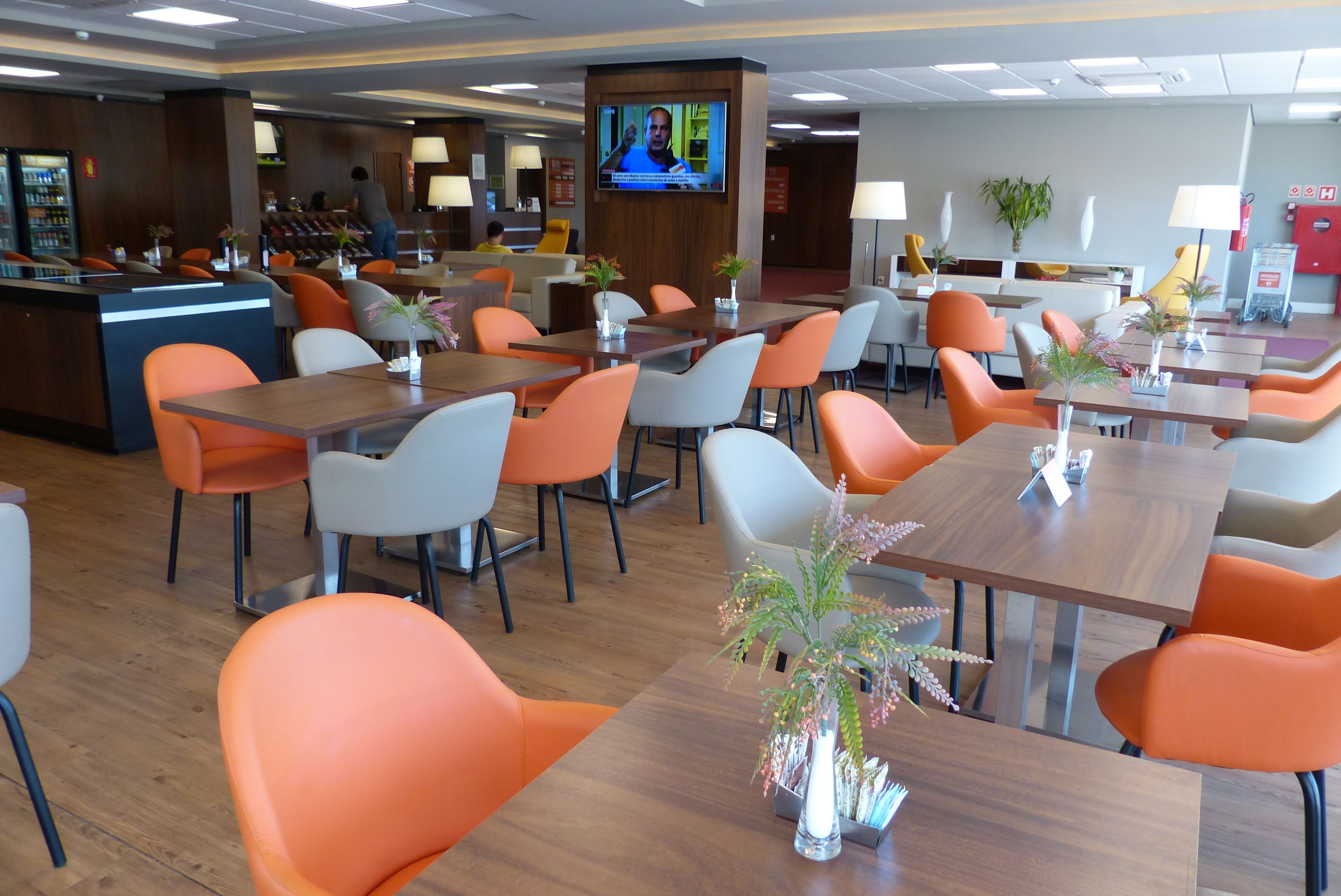 A modern restaurant with orange and white chairs, wooden tables, and a TV screen displaying a news program.